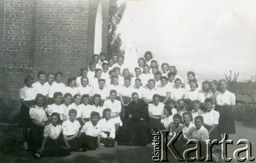 1943-1947, Kidugala, Tanganika.
Ojciec Justynian Maciaszek (1913-1959, proboszcz parafii w Kidugali, współtwórca ośrodka polskiego duszpasterstwa w argentyńskim Martin Coronado) z parafiankami.
Fot. ze zbiorów Archiwum Ojców Franciszkanów w Polskiej Misji Katolickiej w Martin Coronado w Argentynie, akta ojca Justyniana Maciaszka, reprodukcje cyfrowe w  Bibliotece Polskiej im. Ignacego Domeyki w Buenos Aires (Biblioteca Polaca Ignacio Domeyko) i w Ośrodku KARTA w Warszawie
