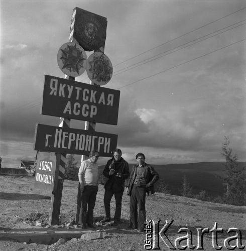 1977, Nieriungri, Jakucka Autonomiczna Socjalistyczna Rosyjska Federacyjna Socjalistyczna Republika Radziecka, ZSRR.
Budowa Bajkalsko-Amurskiej Magistrali Kolejowej (BAM). Operator i dziennikarz Mikołaj Nesterowicz (po prawej) oraz dwaj mężczyźni stoją przy znajdującej się przy wjeździe do miasta tablicy informującej o przynależności administracyjnej terenu.
Fot. Mikołaj Nesterowicz, zbiory Ośrodka KARTA