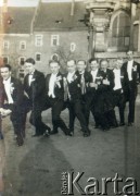 Lata 30., brak miejsca.
Grupa mężczyzn w smokingach pozuje do zdjęcia. W tle widoczny fragment kościoła i kamienice.
Fot. NN, kolekcja Barbary Murzynowskiej, reprodukcje cyfrowe w Ośrodku KARTA