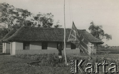 1944, Koja, Uganda.
Mężczyzna wciąga na maszt flagę brytyjską przed budynkiem administracji. W głębi budynek kancelarii i biur. [W osiedlu Koja w latach 1942-1948 mieszkało ok. 3000 Polaków, którzy z żołnierzami Armii Andersa zostali ewakuowani do Iranu].
Fot. NN, zbiory Ośrodka KARTA , udostępniła Maria Wierzchowska [AW - II/1681]