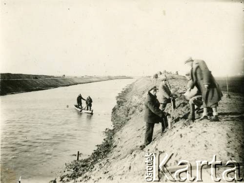 1929-1939, okolice Brześcia nad Bugiem, woj. poleskie, Rzeczpospolita Polska.
Grupa mężczyzn stoi na wale na brzegu rzeki. 1. z prawej widoczny Eugeniusz Lewicki.
Kanał Muchawiecki (Kanał Dniepr-Bug, zwany również Kanałem Królewskim lub Kanałem Rzeczypospolitej) otwarty w 1848 roku kanał żeglowny łączący Dniepr z Wisłą poprzez Pinę (dopływ Prypeci) i Muchawiec (prawostronny dopływ Bugu) stanowiący część szlaku wodnego łączącego Morze Bałtyckie z Morzem Czarnym. Pierwszy odcinek Kanału zbudowano w latach 1775-1784, następnie, w latach 1846-1848, Kanał był rozbudowywany przez władze rosyjskie. W latach 1929-1939, w czasach administracji II Rzeczypospolitej, poddawany gruntowym pracom remontowym. 
Fot. NN, kolekcja Eugeniusza Lewickiego, zbiory Ośrodka KARTA