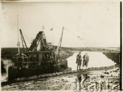 1929-1939, okolice Brześcia nad Bugiem, woj. poleskie, Rzeczpospolita Polska.
Pogłębiarka podczas pracy. Na brzegu rzeki widoczni dwaj mężczyźni, 1. z prawej Eugeniusz Lewicki, obok niego nadzorujący pracę urzędnik państwowy.
Kanał Muchawiecki (Kanał Dniepr-Bug, zwany również Kanałem Królewskim lub Kanałem Rzeczypospolitej) otwarty w 1848 roku kanał żeglowny łączący Dniepr z Wisłą poprzez Pinę (dopływ Prypeci) i Muchawiec (prawostronny dopływ Bugu) stanowiący część szlaku wodnego łączącego Morze Bałtyckie z Morzem Czarnym. Pierwszy odcinek Kanału zbudowano w latach 1775-1784, następnie, w latach 1846-1848, Kanał był rozbudowywany przez władze rosyjskie. W latach 1929-1939, w czasach administracji II Rzeczypospolitej, poddawany gruntowym pracom remontowym. 
Fot. NN, kolekcja Eugeniusza Lewickiego, zbiory Ośrodka KARTA