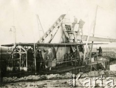 1929-1939, okolice Brześcia nad Bugiem, woj. poleskie, Rzeczpospolita Polska.
Pogłębiarka podczas pracy. Na pokładzie jednostki pośród mężczyzn widoczny Eugeniusz Lewicki (3. z lewej). Obok niego, w kożuchu oraz czapce z orzełkiem, nadzorujacy pracę urzędnik.
Kanał Muchawiecki (Kanał Dniepr-Bug, zwany również Kanałem Królewskim lub Kanałem Rzeczypospolitej) otwarty w 1848 roku kanał żeglowny łączący Dniepr z Wisłą poprzez Pinę (dopływ Prypeci) i Muchawiec (prawostronny dopływ Bugu) stanowiący część szlaku wodnego łączącego Morze Bałtyckie z Morzem Czarnym. Pierwszy odcinek Kanału zbudowano w latach 1775-1784, następnie, w latach 1846-1848, Kanał był rozbudowywany przez władze rosyjskie. W latach 1929-1939, w czasach administracji II Rzeczypospolitej, poddawany gruntowym pracom remontowym. 
Fot. NN, kolekcja Eugeniusza Lewickiego, zbiory Ośrodka KARTA