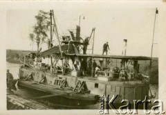 1929-1939, okolice Brześcia nad Bugiem, woj. poleskie, Rzeczpospolita Polska.
Pogłębiarka podczas pracy. Kanał Muchawiecki (Kanał Dniepr-Bug, zwany również Kanałem Królewskim lub Kanałem Rzeczypospolitej) otwarty w 1848 roku kanał żeglowny łączący Dniepr z Wisłą poprzez Pinę (dopływ Prypeci) i Muchawiec (prawostronny dopływ Bugu) stanowiący część szlaku wodnego łączącego Morze Bałtyckie z Morzem Czarnym. Pierwszy odcinek Kanału zbudowano w latach 1775-1784, następnie, w latach 1846-1848, Kanał był rozbudowywany przez władze rosyjskie. W latach 1929-1939, w czasach administracji II Rzeczypospolitej, poddawany gruntowym pracom remontowym. 
Fot. NN, kolekcja Eugeniusza Lewickiego, zbiory Ośrodka KARTA