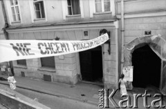 Po 28.05.1981, Kraków, Polska.
Ulica Gołębia. Studentka czyta ogłoszenie z informacją o śmierci kardynała Stefana Wyszyńskiego wywieszone przy wejściu do kamienicy pod numerem 6. Między budynkami rozpięty transparent z hasłem: 