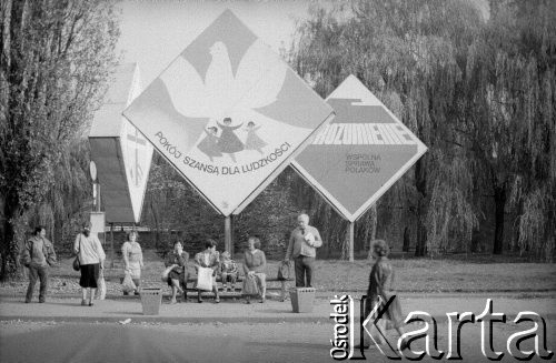 1989, Kraków, Polska.
Ludzie czekający na przystanku autobusowym. Na skwerze konstrukcje z hasłami propagandowymi 