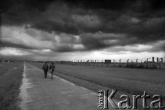 1986, Oświęcim, Polska.
KL Auschwitz Birkenau. Droga do obozu. Na drodze kobieta i mężczyzna. Z prawej ogrodzenie i ruiny budynków obozowych. W głębi wartownia i brama główna. 
Fot. Piotr Dylik, zbiory Ośrodka KARTA