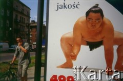 Lata 90., Kraków, Polska.
Plakat reklamowy przedstawiający zawodnika sumo. Z lewej przechodzień prowadzi rower.
Fot. Piotr Dylik, zbiory Ośrodka KARTA