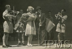 1942, Teheran, Iran.
Występ w obozie dla polskich uchodźców, aktorzy na scenie. Obok aktorki z bukietem kwiatów stoi sobowtór Charliego Chaplina, w tle orkiestra.
Fot. NN, Instytut Polski i Muzeum im. gen. Sikorskiego w Londynie [album Ludwika Naimskiego].