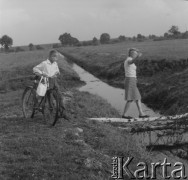 Lata 60. lub 70., Górecko Kościelne, woj. lubelskie, Polska.
Chłopiec z rowerem patrzy na kobietę przechodzącą po gałęziach przerzuconych nad strumykiem, który  płynie między polami.
Fot. Jerzy Konrad Maciejewski, zbiory Ośrodka KARTA