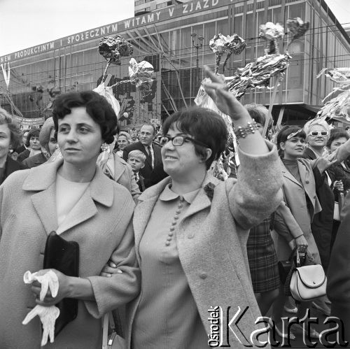 01.05.1968, Warszawa, Polska.
Pochód pierwszomajowy w centrum Warszawy. Dwie kobiety spoglądające w dal, z tyłu wiwatujący tłum ludzi z kwiatami z folii aluminiowej i innymi stroikami. W tle Domy Towarowe 